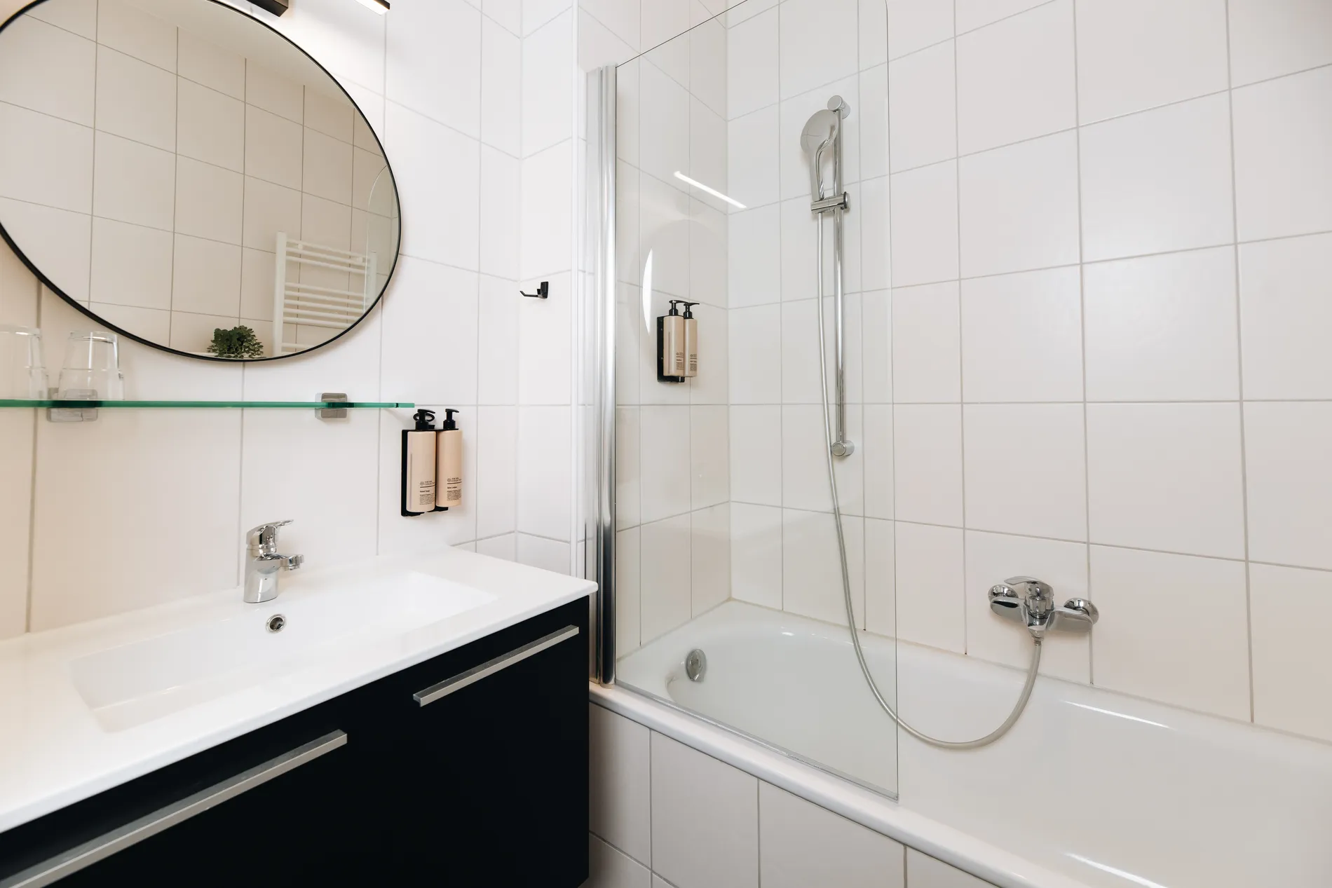 Modern badkamerinterieur met zwart wastafelmeubel, ronde spiegel en een bad-douchecombinatie met glazen wand.